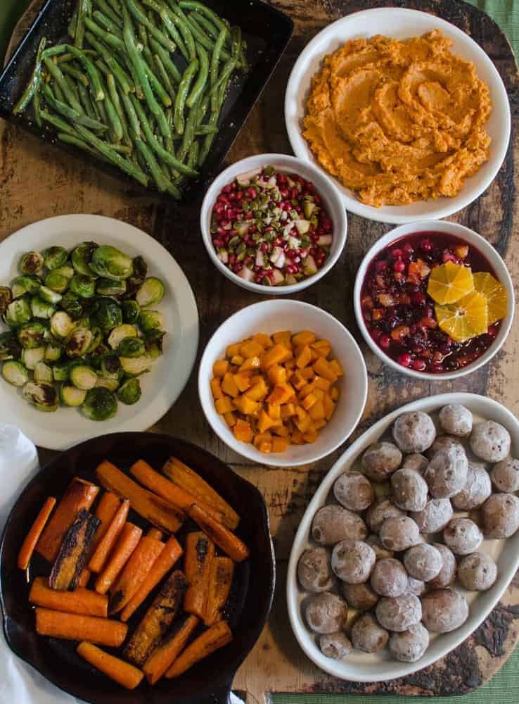 An overhead view of various vegetable dishes, including roasted green beans, mashed sweet potatoes, Brussels sprouts, diced squash, cranberry relish with orange, a cucumber salad with pomegranate, roasted carrots, and baby potatoes.