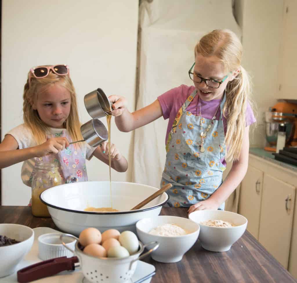 Two young girls wearing aprons pour ingredients into a large mixing bowl on a kitchen table, preparing to bake applesauce oatmeal cookies. Bowls of eggs, flour, and other baking supplies surround them as they carefully measure and mix.