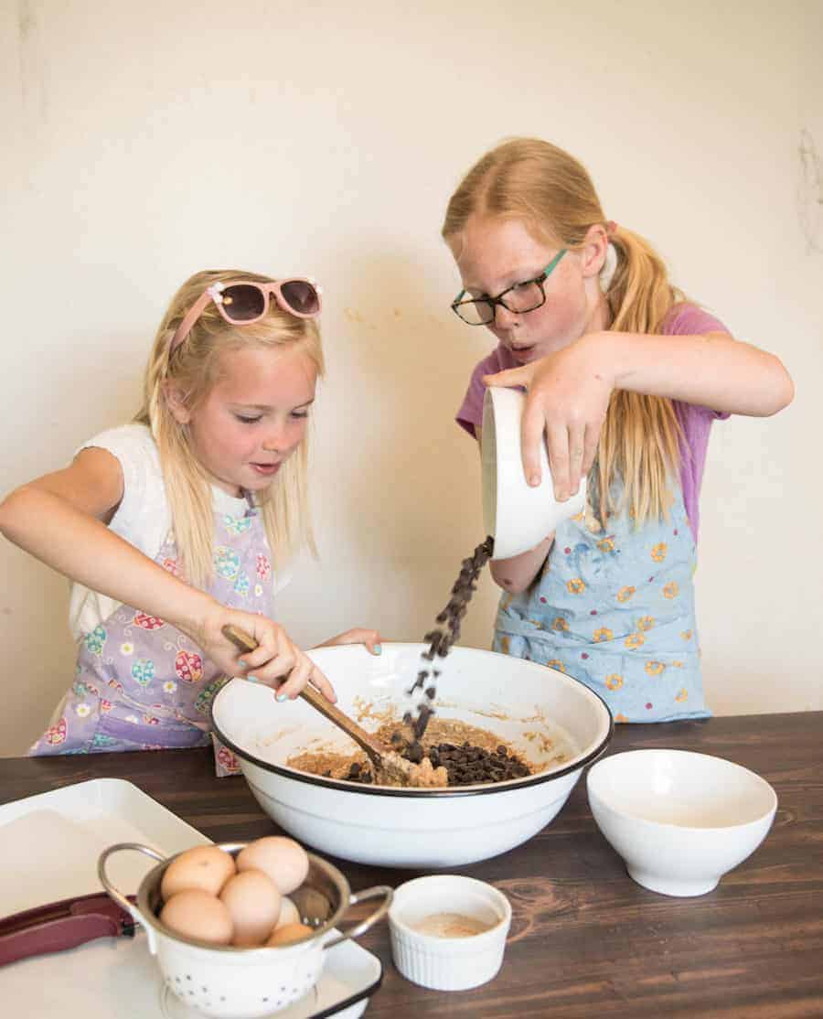 Two kids making Healthy Applesauce Cookies where one is mixing the dough and the other is pouring in the chocolate chips from a small white bowl.