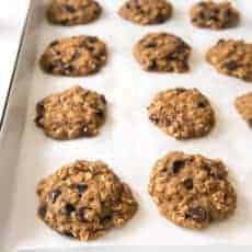 A baking sheet lined with parchment paper holds several freshly baked applesauce oatmeal cookies with chocolate chips arranged in rows.