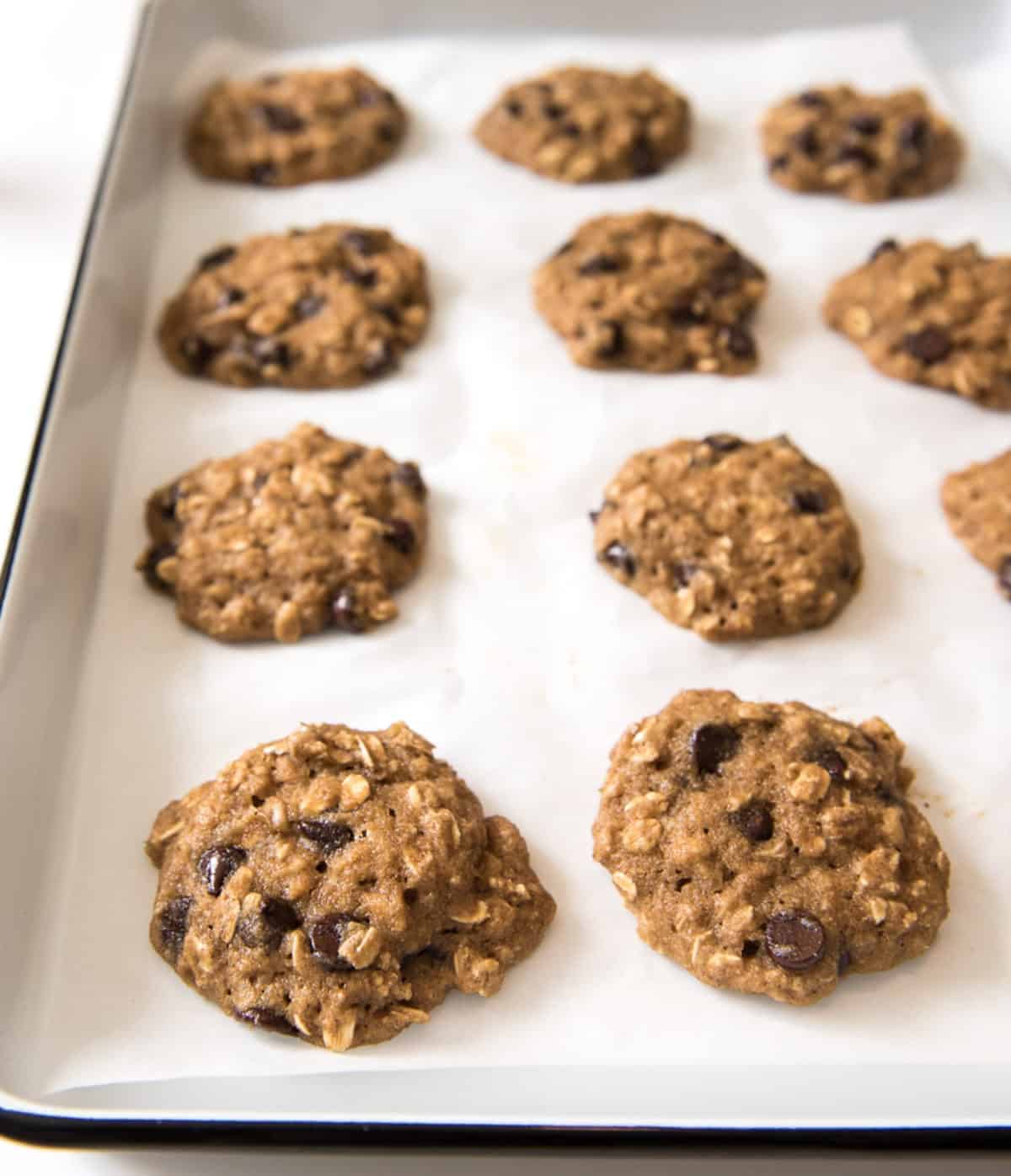 A dozen baked Healthy Applesauce Cookies on white parchment paper on a white enamelware baking sheet with a black rim.