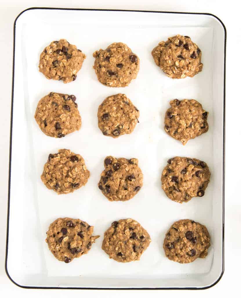 A white baking tray holds twelve evenly spaced, unbaked applesauce oatmeal cookie dough balls arranged in rows, ready for baking.