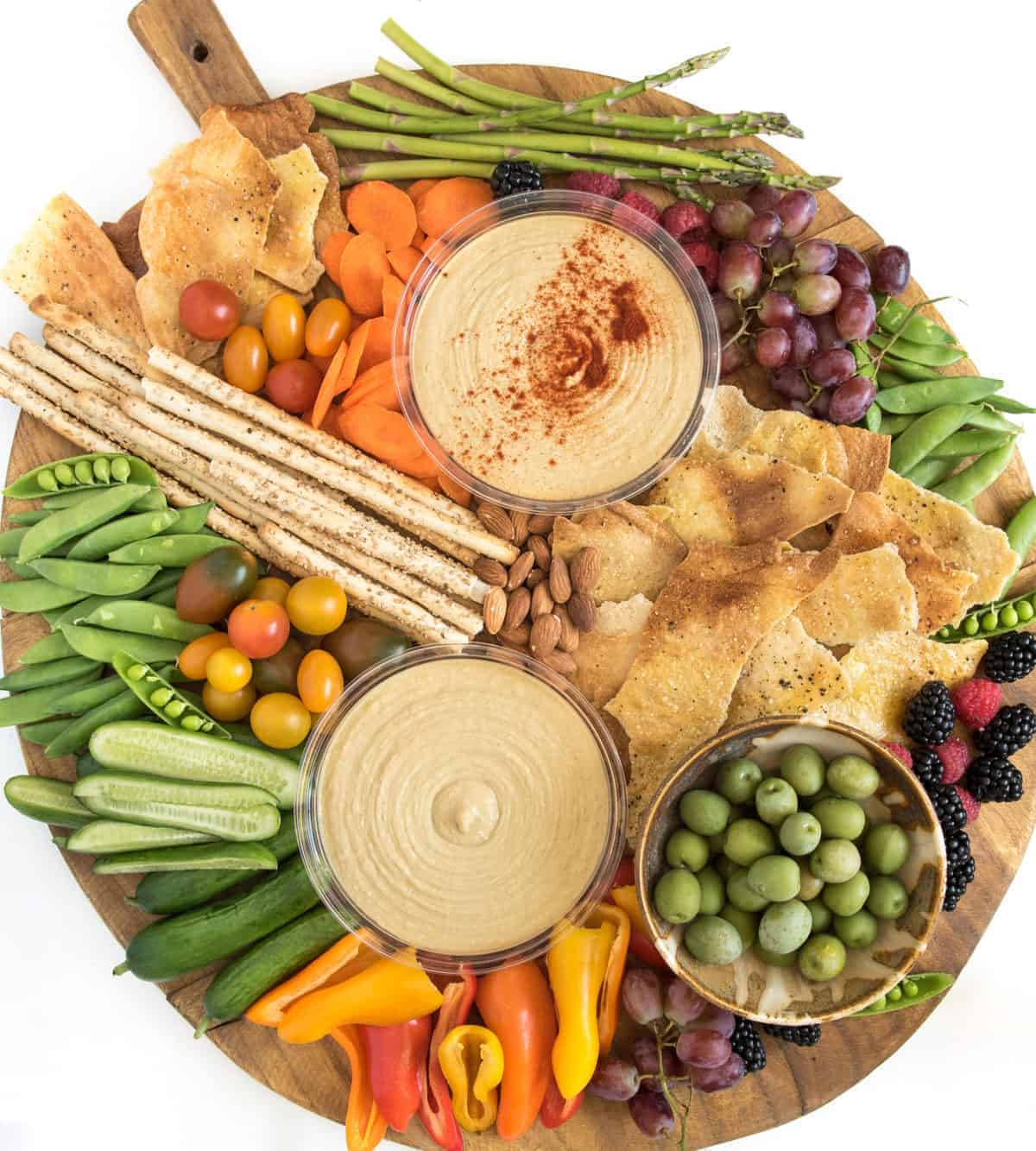 A simple DIY hummus platter is a beautiful addition to any party, or it can be a meal all on it's own! A round wooden hummus platter topped with two bowls of hummus, a bowl of green olives, crackers, breadsticks, cherry tomatoes, snap peas, cucumber slices, mini bell peppers, asparagus, grapes, almonds, blackberries, and carrots.