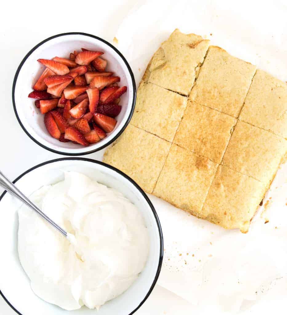 A baked cake cut into squares sits on parchment paper, resembling the best strawberry shortcake. Accompanying it are two bowls: one filled with sliced strawberries and the other with whipped cream. A silver spoon rests invitingly in the whipped cream bowl.