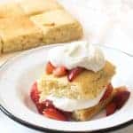A plate features an enticing serving from an easy strawberry shortcake recipe: a slice of sponge cake adorned with fresh, sliced strawberries and a generous dollop of whipped cream. More cake awaits in the background, tempting you for seconds.