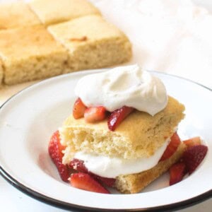 A plate features an enticing serving from an easy strawberry shortcake recipe: a slice of sponge cake adorned with fresh, sliced strawberries and a generous dollop of whipped cream. More cake awaits in the background, tempting you for seconds.