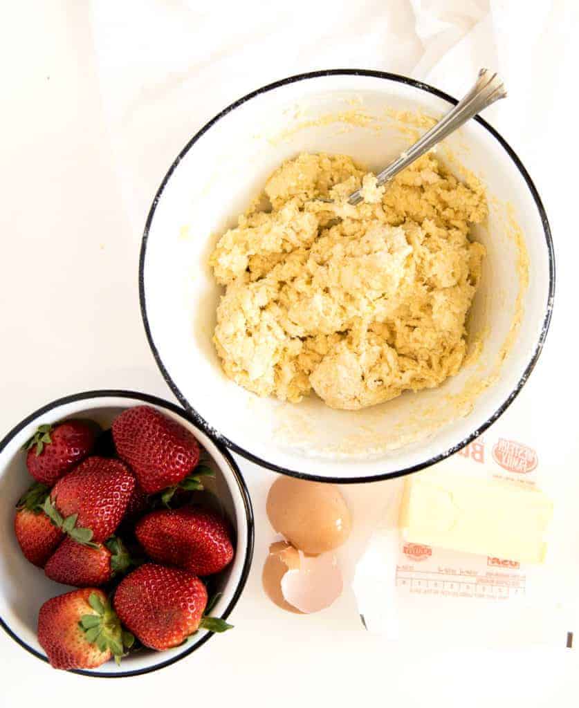 A bowl of cookie dough with a spoon, fresh strawberries, two brown eggs (one cracked), and a stick of butter on a white surface&mdash;perfect ingredients for an easy strawberry shortcake recipe.