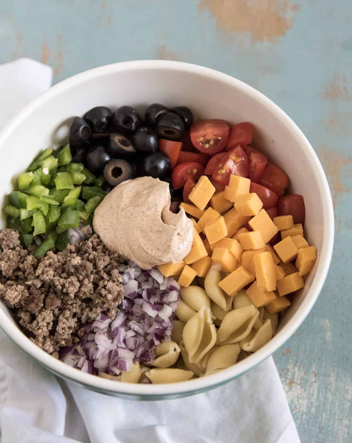 Creamy taco pasta salad made with pasta, hamburger, chopped vegetables, and a creamy taco inspired dressing to bring it all together. Bonus: It's done in about 20 minutes! Ingredients for Creamy Taco Pasta Salad in a white bowl, including ground beef, bell peppers, black olives, grape tomatoes, cheddar cheese, shell pasta, red onions, and a dollop of the dressing.