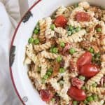 A white round serving bowl with chicken, bacon ranch pasta salad with halved grape tomatoes and green peas