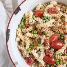 A white round serving bowl with chicken, bacon ranch pasta salad with halved grape tomatoes and green peas