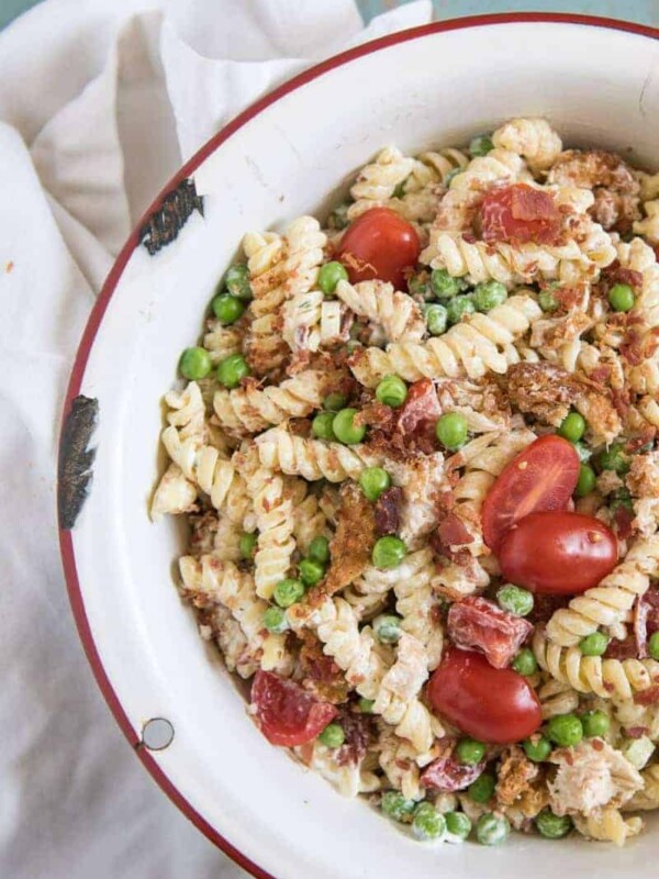 A white round serving bowl with chicken, bacon ranch pasta salad with halved grape tomatoes and green peas