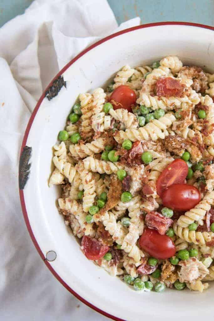 A white round serving bowl with chicken, bacon ranch pasta salad with halved grape tomatoes and green peas