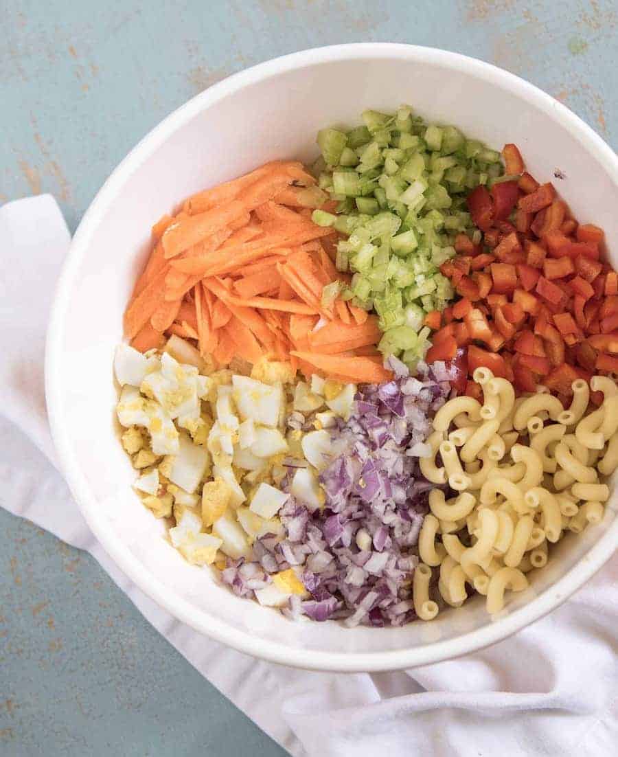 Ingredients for Classic Macaroni Salad in a white bowl including grated carrot, cooked macaroni pasta, and chopped red bell pepper, celery, red onion, and hard boiled eggs.