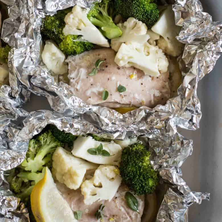 Two foil packets with salmon, broccoli, cauliflower, and a lemon wedge