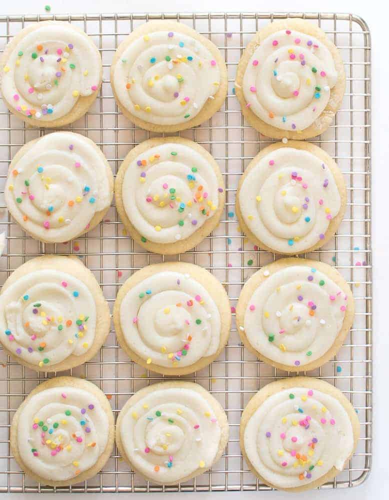 pale cookies on a wire rack with white frosting and colored sprinkles on top.