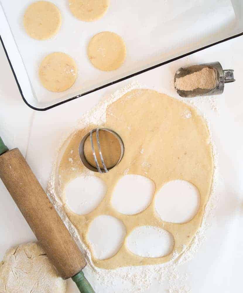 cookie dough rolled out with cookie cutter, flour scoop, and wooden rolling pin next to a white cookie sheet with cut out cookies on it.