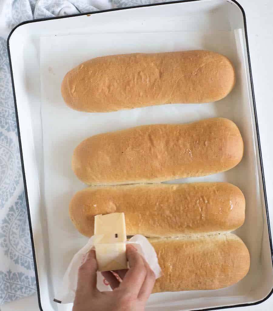 four long rolls of bread lightly brown on top sitting in a white pan with hand rubbing butter on them.