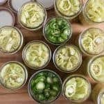 A top view showcases how to pickle peppers with an array of jars filled with vibrant pickled peppers. Most jars contain light yellow pepper rings, while two have green jalapeño slices. The closely arranged jars rest on a wooden surface, their lids adding a rustic touch in the background.