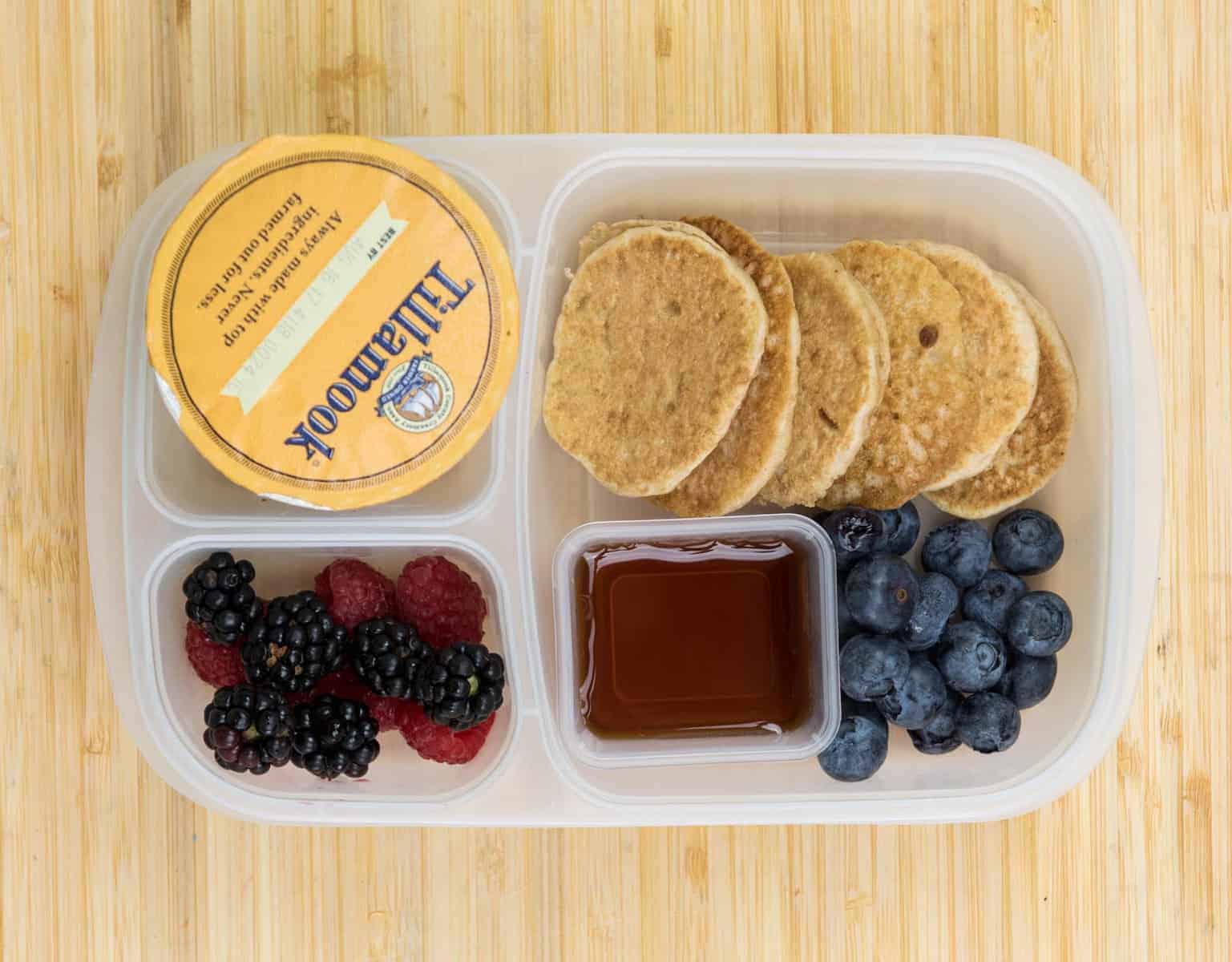 Lunch Box Ideas - Breakfast for Lunch Box Top view of a lunch box with Mini whole wheat pancakes, Maple syrup, Blackberries + raspberries + blueberries, and Yogurt.