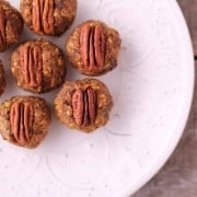 A white plate with seven pumpkin date balls topped with pecan halves, arranged on a rustic wooden surface. The snacks boast a textured, nutty exterior.