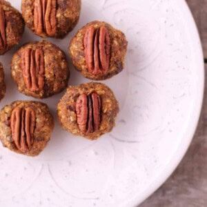 A white plate with seven pumpkin date balls topped with pecan halves, arranged on a rustic wooden surface. The snacks boast a textured, nutty exterior.