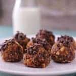 A plate of six chocolate chip energy bites is placed on a light blue surface. The no-bake pumpkin balls are sprinkled with mini chocolate chips. A small glass bottle of milk is blurred in the background, adding a cozy touch to this delightful scene.
