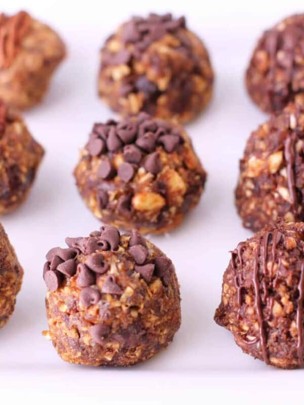 A plate with nine no bake pumpkin balls arranged in rows. Each ball is topped differently: some with pecans, some with chocolate chips, and others drizzled with chocolate. They have a textured, nutty appearance.
