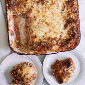 A large baking dish of cheesy, make-ahead lasagna with a corner piece missing. Two servings of lasagna are placed on white plates beside the dish. The lasagna has a golden, slightly browned top.