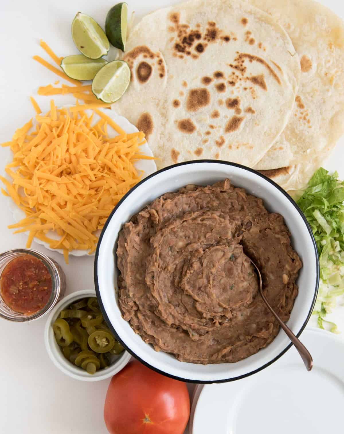 Slow cooker refried beans are the simplest way to make rich and hearty refried beans at home - they are worlds above their canned counterparts. Refried beans in a white enamel bowl with a spoon in it surrounded by taco fixings, including tortillas, shredded lettuce, shredded cheddar cheese, salsa, sliced jalapeños and a tomato.