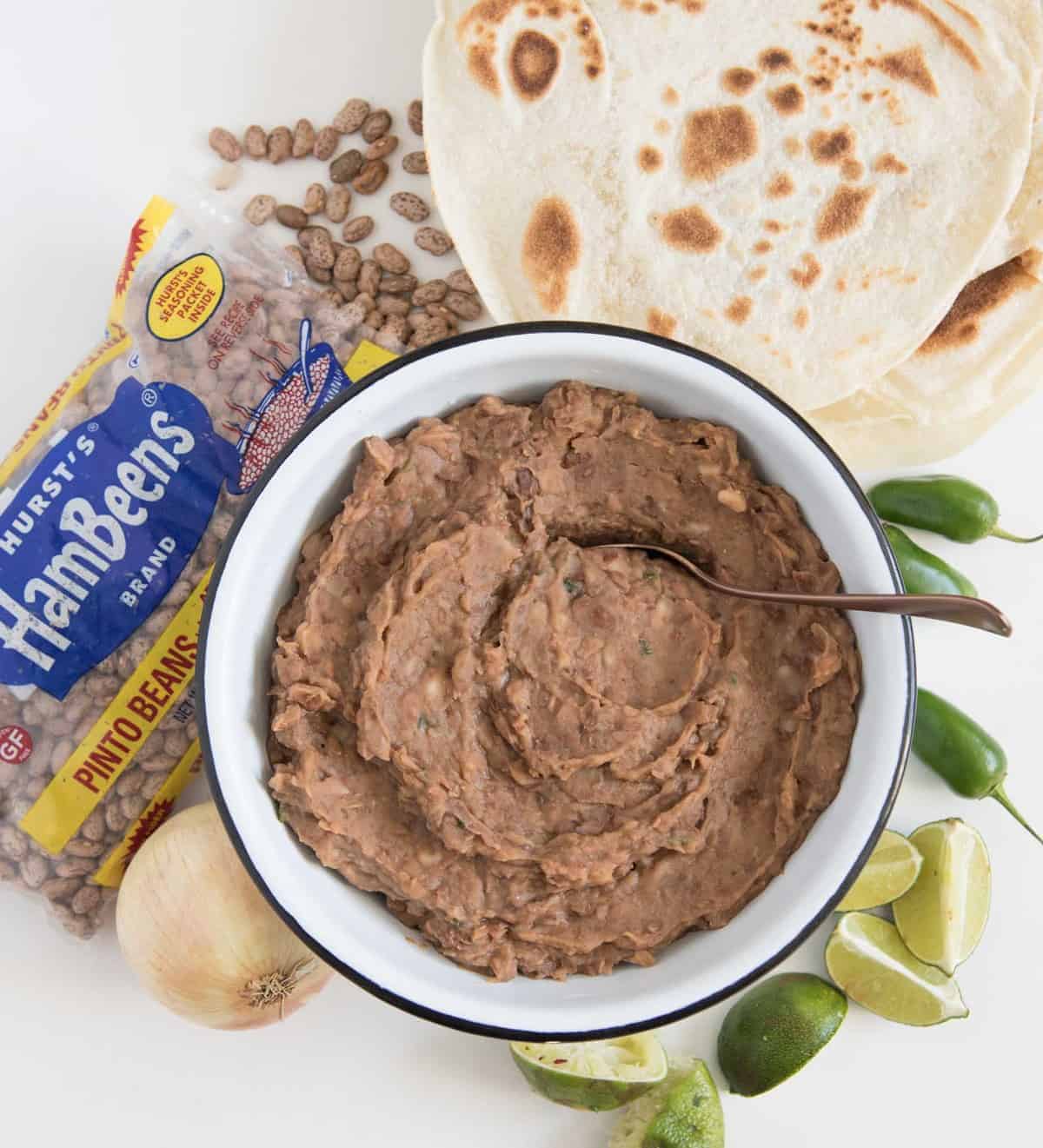 Slow cooker refried beans are the simplest way to make rich and hearty refried beans at home. Refried beans in a white bowl with spoon in it and a bag of pinto beans, onion, some lime slices, tortillas, and whole jalapeños next to it.
