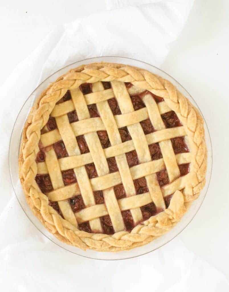 A golden-brown cherry pie with a lattice crust and braided edge sits on a clear glass pie dish atop a white cloth, reminiscent of the classic charm found in a homemade coconut cream pie.