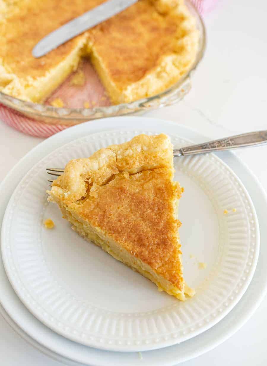 A piece of old fashioned pie is on a white salad plate on top of a plain white plate with a silver fork on the plate. The rest of the pie is pictured in the background in a clear pie dish with a silver butter knife on it.