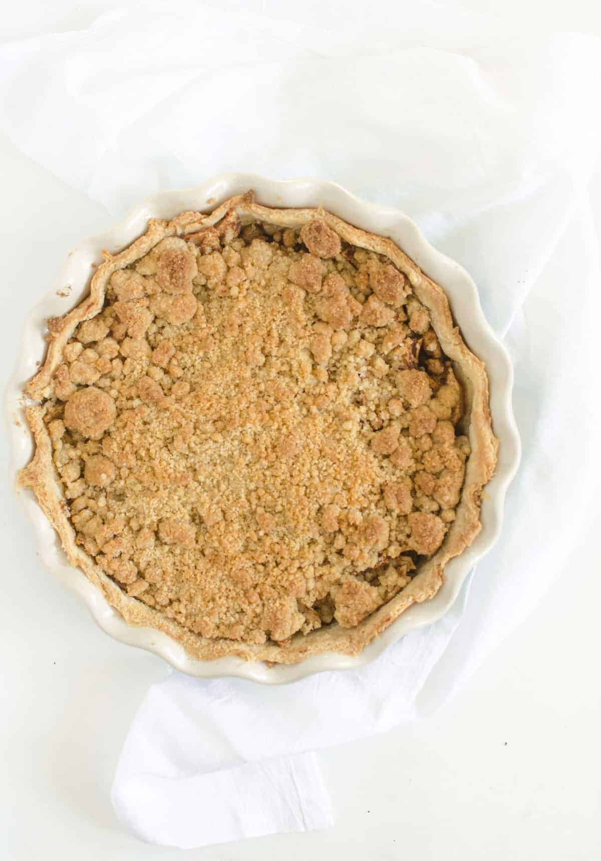 A round dutch apple pie with a golden brown crumb topping sits in a scalloped-edge pie dish on a white cloth and white background.