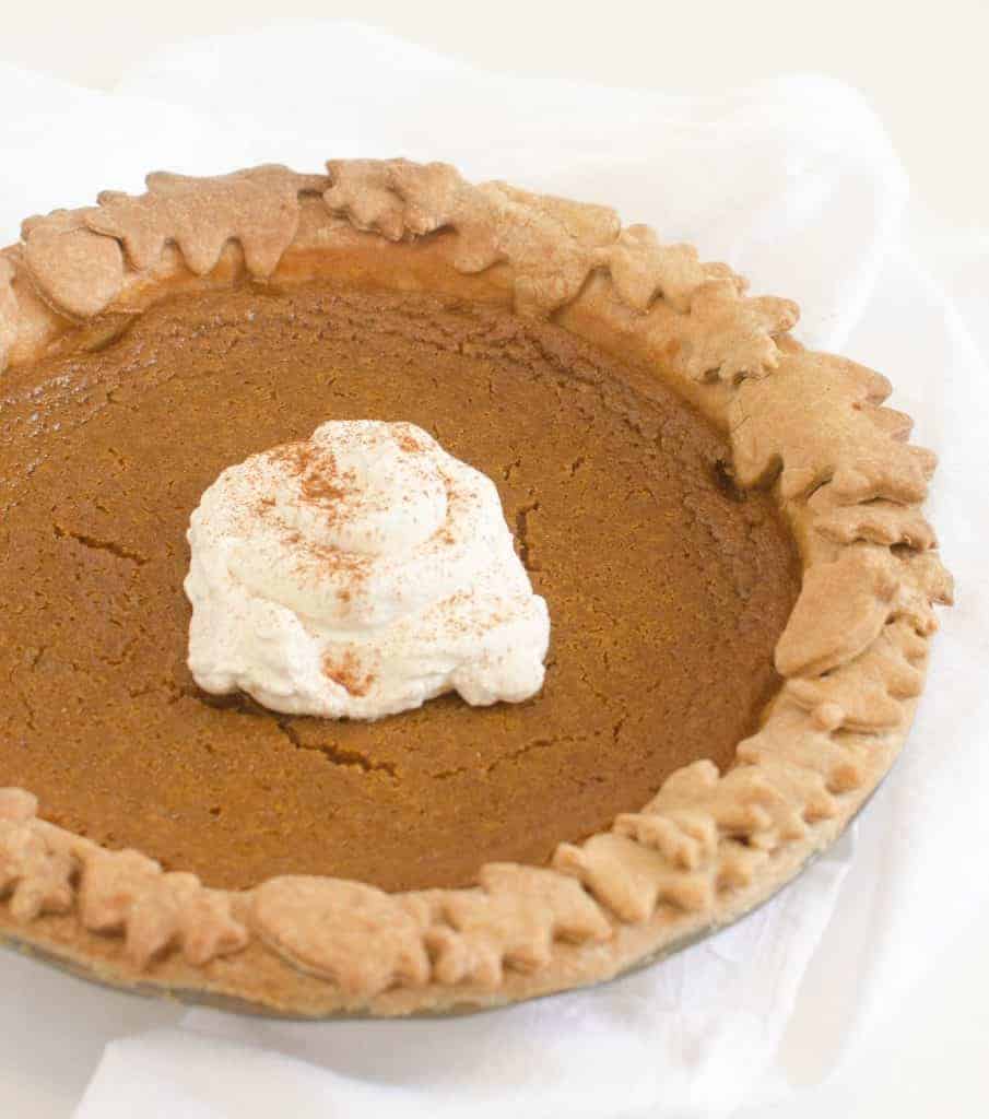 A pumpkin pie with decorative leaf-shaped crust edges, topped with a dollop of whipped cream reminiscent of a coconut cream pie, is sprinkled with cinnamon and sits on a white cloth background.