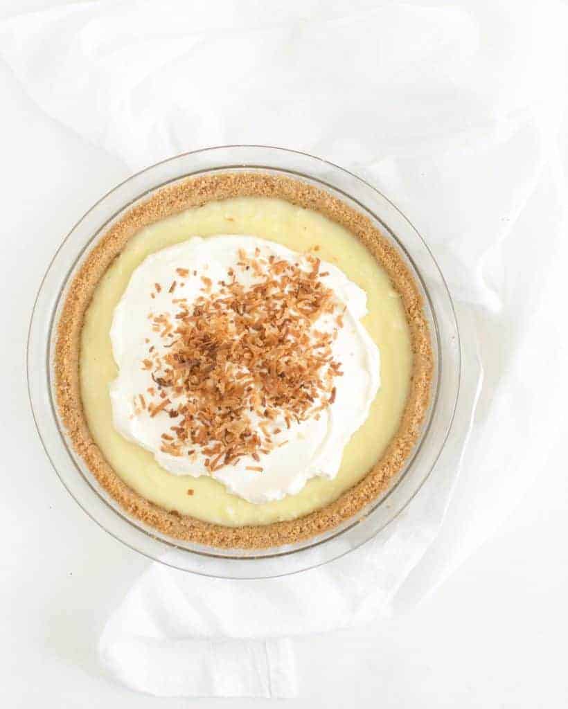 A classic coconut cream pie with a graham cracker crust, topped with whipped cream and toasted shredded coconut, sits on a clear glass pie dish against a white background.