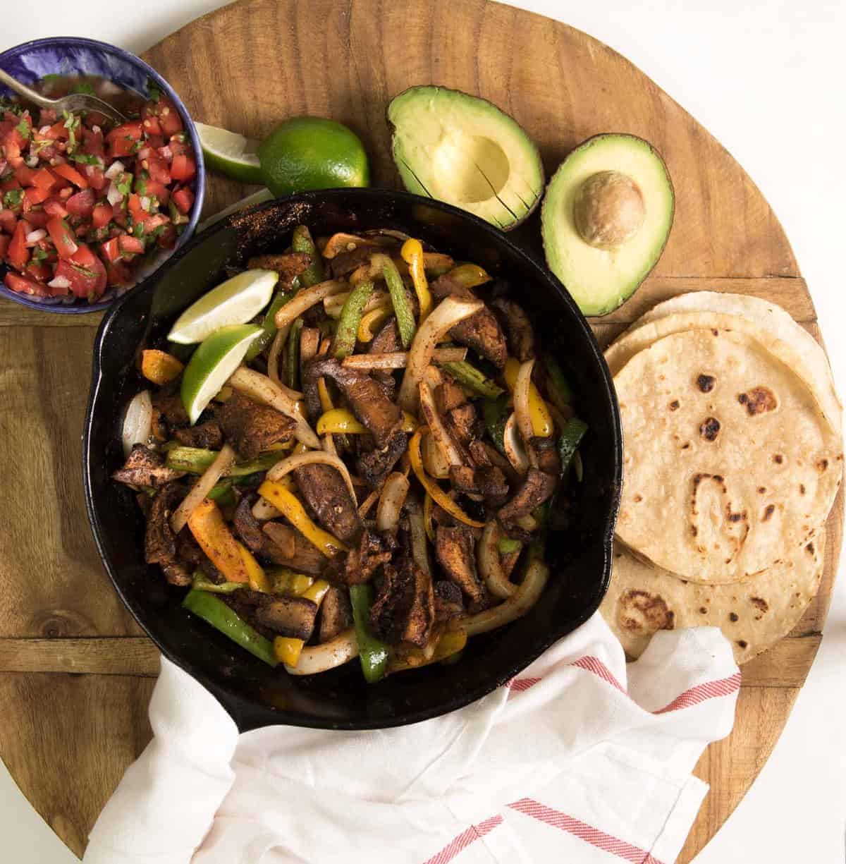 tortillas, salsa, avocado, and lime on a wooden cutting board with the taco filling in a cast iron pan