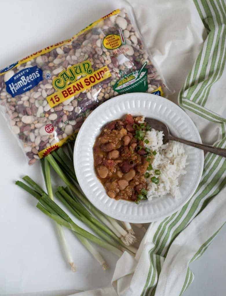 beans and rice in a white bowl with a bags of hurst's cajun 15 bean soup bean mix and green onions on the side