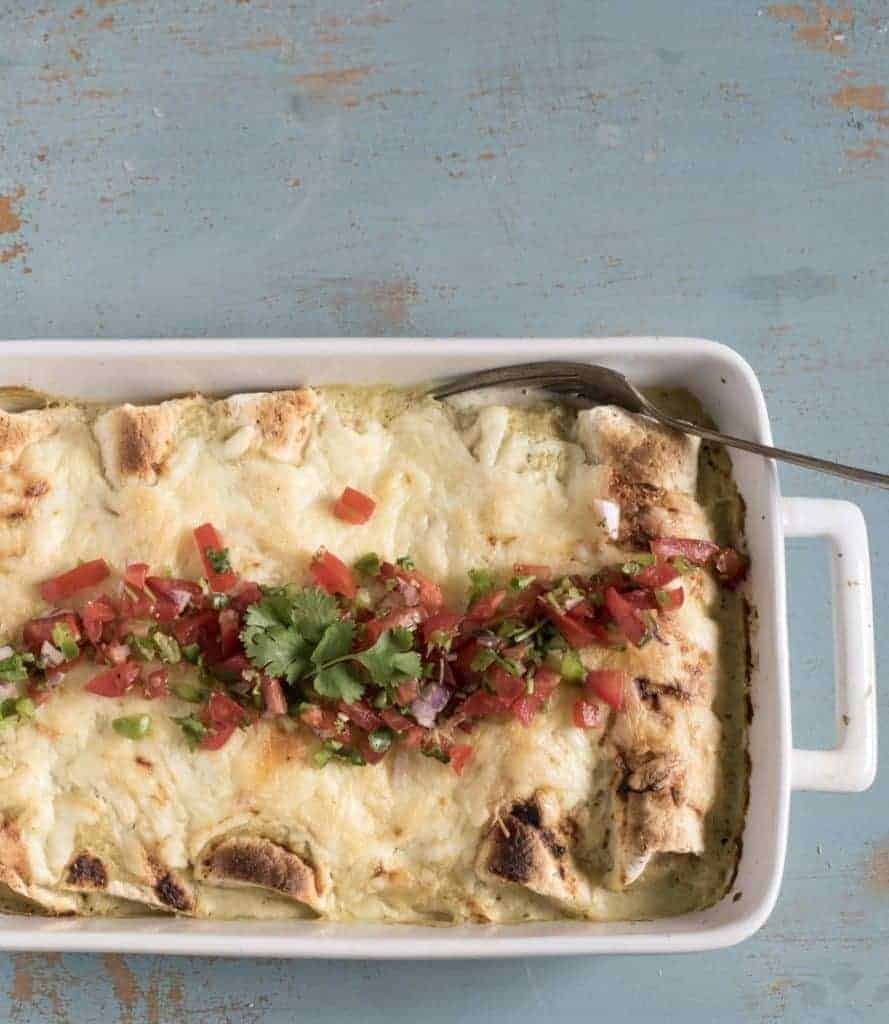 A white rectangular baking dish filled with baked enchiladas topped with melted cheese, pico de gallo and fresh cilantro