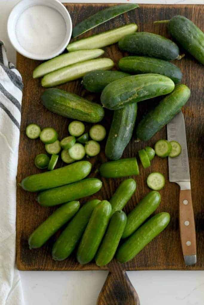 A wooden cutting board with whole mini cucumbers, sliced mini cucumbers, and spears of mini cucumbers with a knife and a small bowl of salt