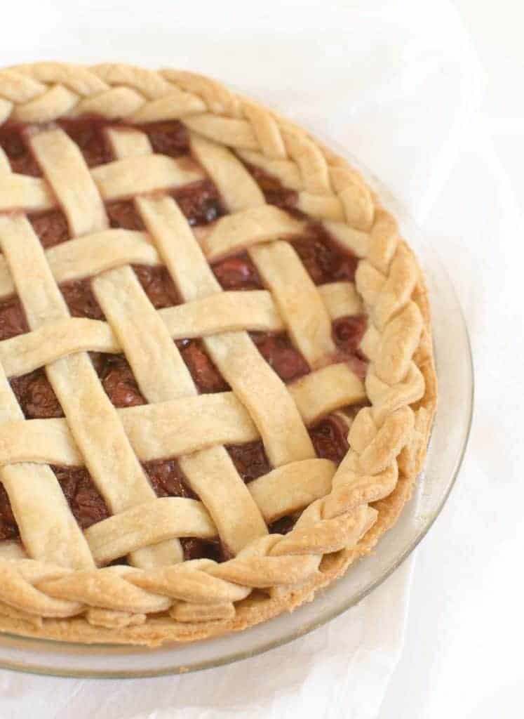 A baked pie with a golden-brown lattice crust and braided edge, filled with dark red fruit filling, displayed in a glass pie dish on a white background—a delicious contrast to any Flourless Chocolate Torte Recipe.