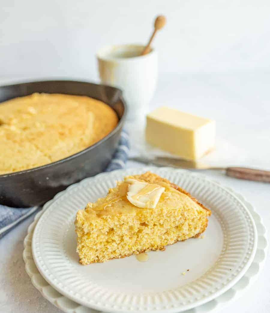 slice of cornbread with butter and honey on top with pan of cornbread behind it