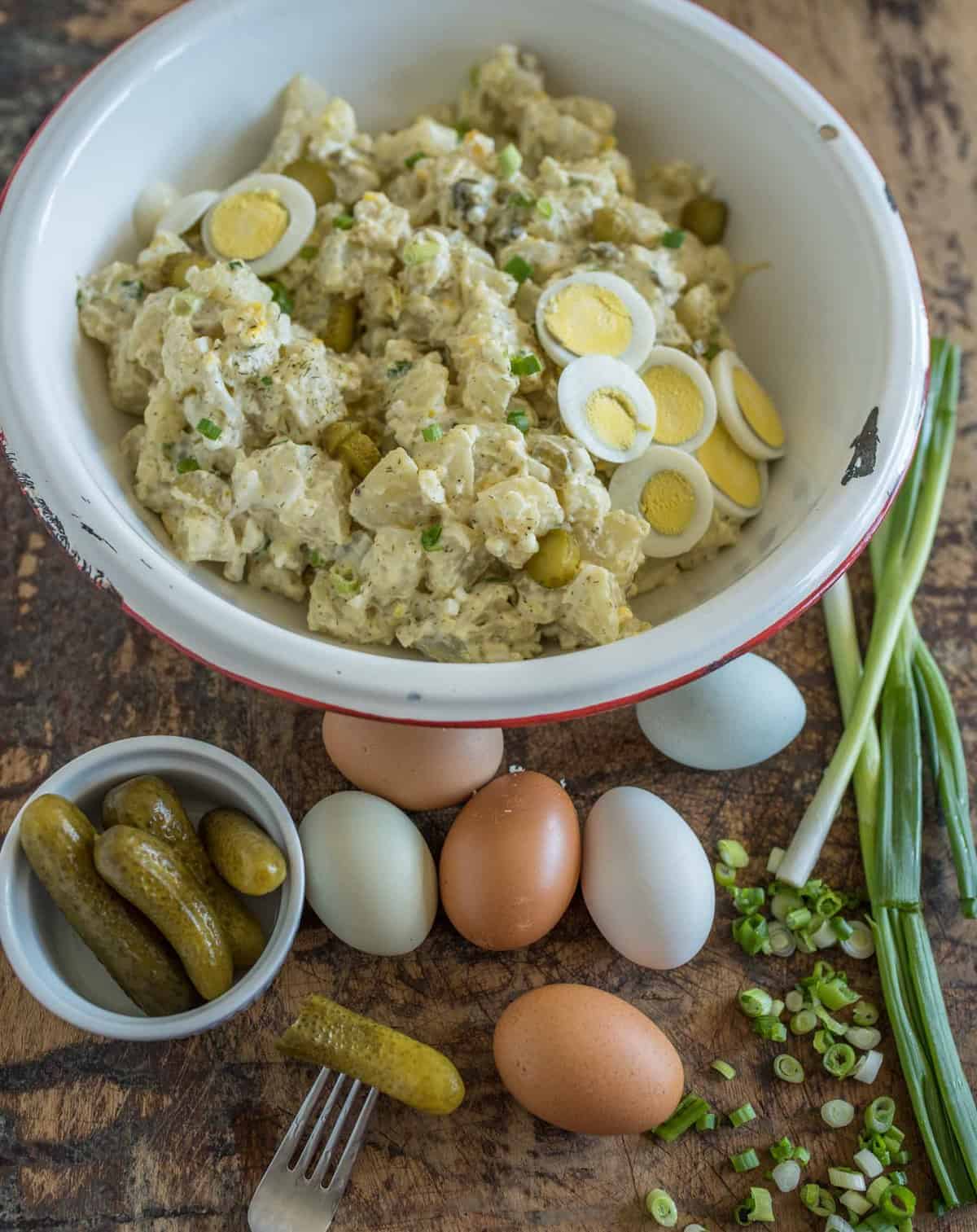 cut up potatoes and eggs in a white bowl with small pickles, eggs, and green onions beside the bowl.