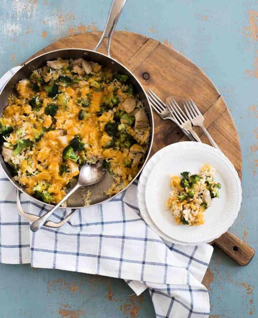 A skillet with chicken broccoli rice casserole with melted cheese and a white round plate with a serving of chicken broccoli rice casserole on a wooden board with a plaid cloth