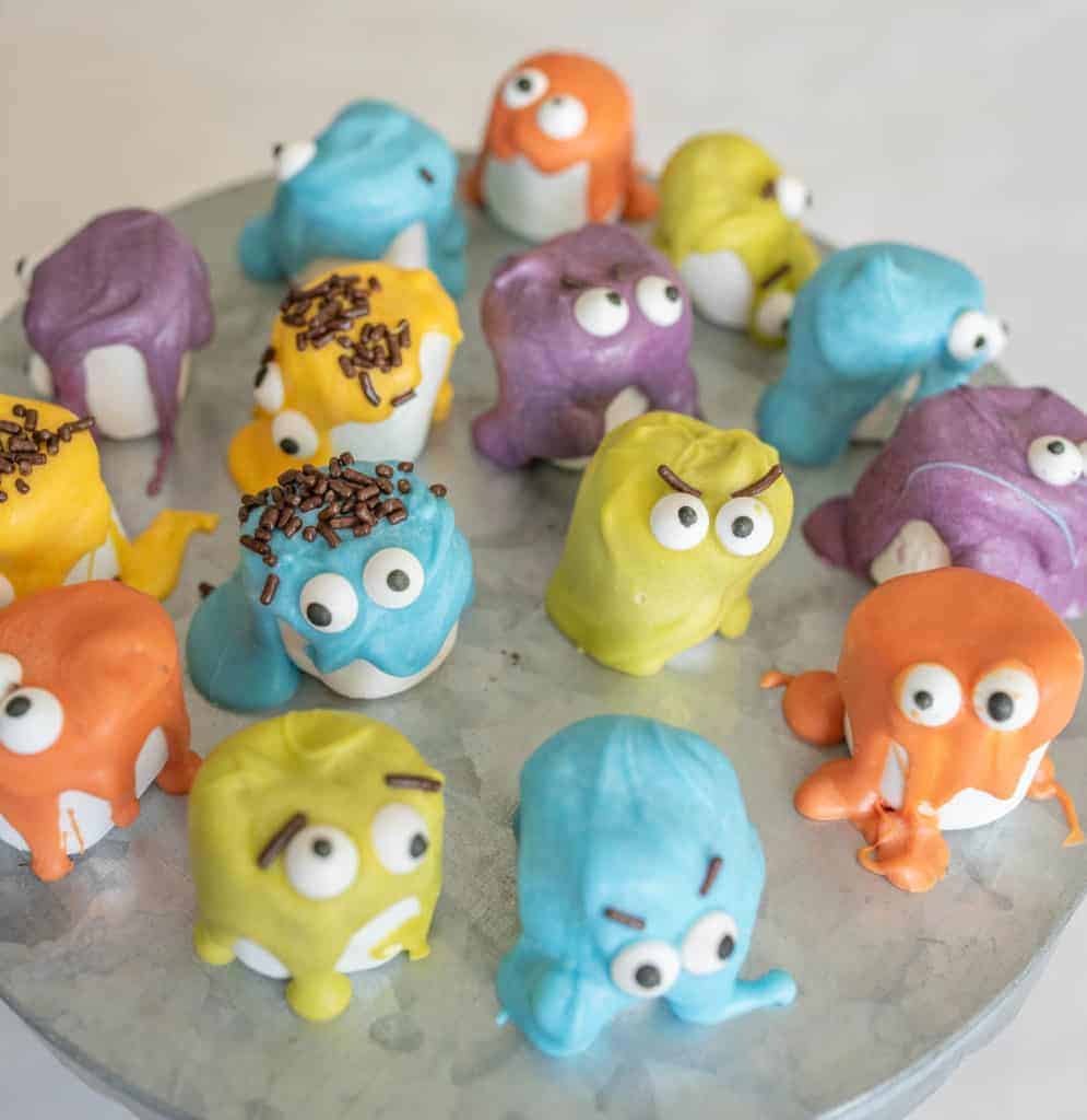 Colorful cake balls decorated to look like whimsical monster creatures with big candy eyes, expressive eyebrows, and bright icing—perfect lunch box ideas—displayed on a round tray in orange, blue, yellow, purple, and green.