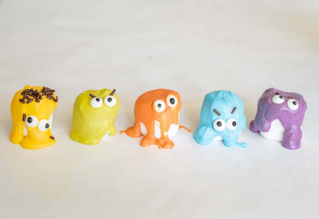 Five colorful, cartoonish monster-shaped treats with big eyes and playful expressions are lined up on a white surface—perfect for creative lunch box ideas. The monsters come in yellow, green, orange, blue, and purple.