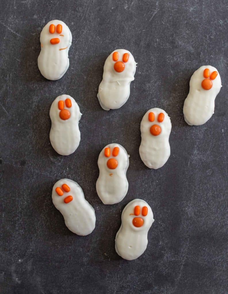 Eight cookie sandwiches coated in white chocolate to resemble ghosts are arranged on a dark surface—perfect for lunch box ideas. Each ghost has two small orange oval eyes and a round orange nose made of candy or icing.