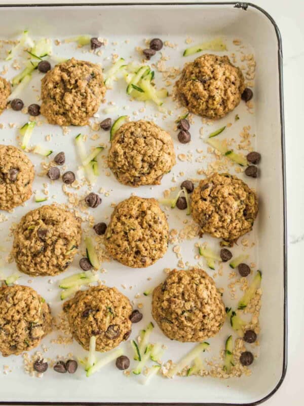 A baking tray filled with freshly baked zucchini cookies with oatmeal and chocolate chips sits surrounded by scattered raw oats, chocolate chips, and zucchini strips, enhancing the fresh, homemade aesthetic.