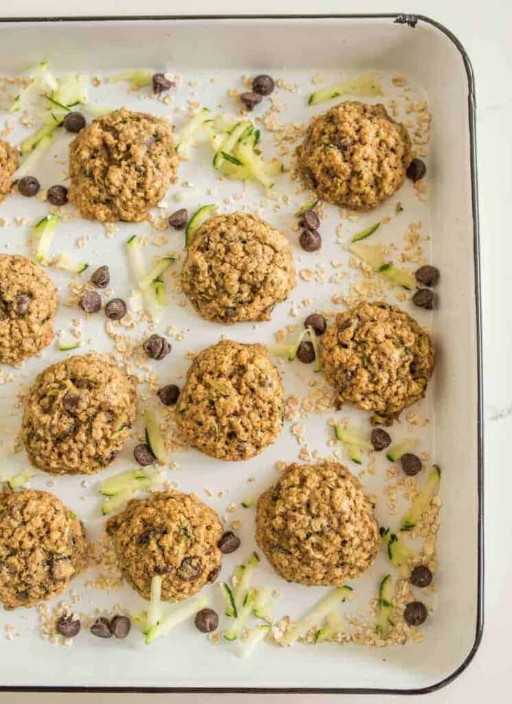 A baking tray filled with freshly baked zucchini cookies with oatmeal and chocolate chips sits surrounded by scattered raw oats, chocolate chips, and zucchini strips, enhancing the fresh, homemade aesthetic.