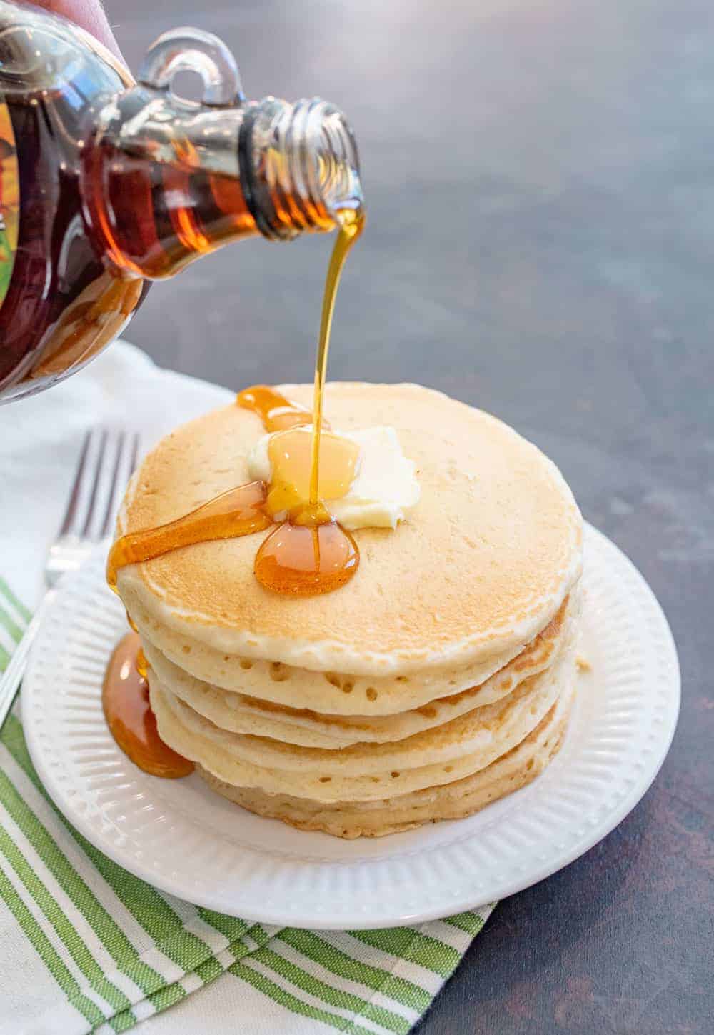 stack of pancakes on a white plate with syrup being poured over on a white and green towel.