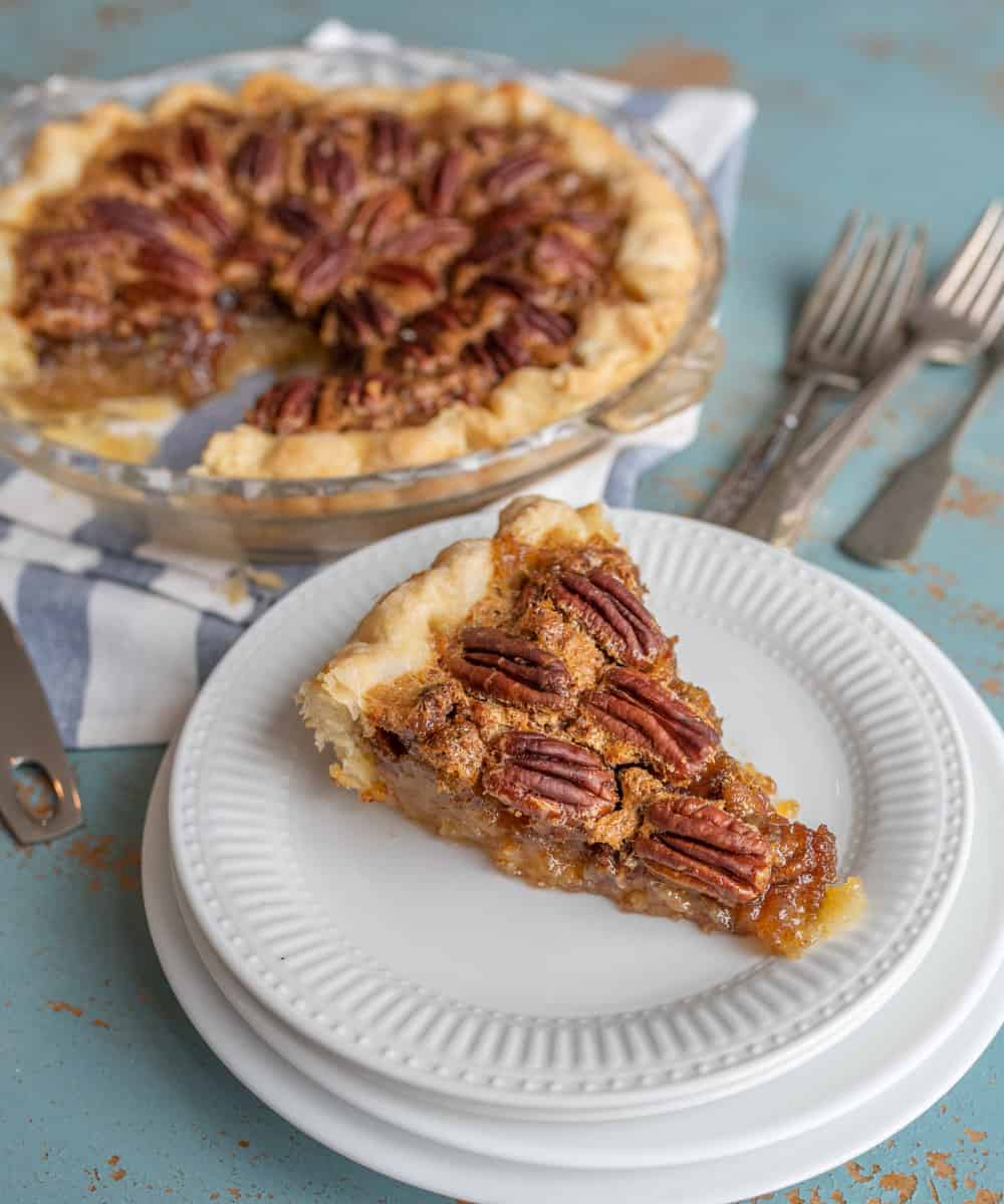 In the foreground, a piece of pecan pie is on a fancy, white dessert plate. Under the dessert plate are other bigger, plain white plates. Above the piece of pie to the right are some silver forks. In the upper left-hand side in the background, is the rest of the pie in a clear pie pan on top of a blue and white striped kitchen towel.